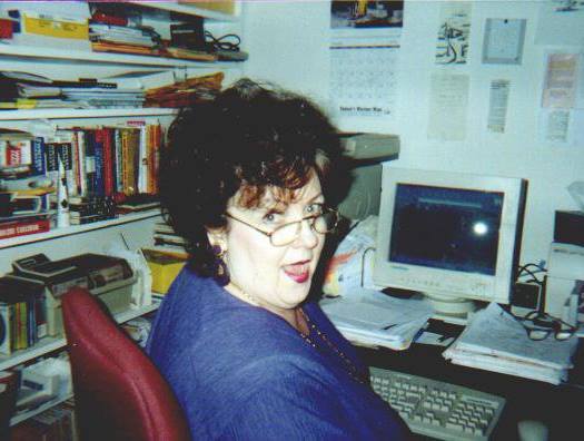 A white woman with dark curly short hair and glasses is shown seated at a computer desk with clutter bookshelves. There's an old computer with a small monitor in front of her as well as stacks of papers and an extra pair of glasses. She's wearing a blue top and is turned halfway to face the camera with a silly, joyful expression.