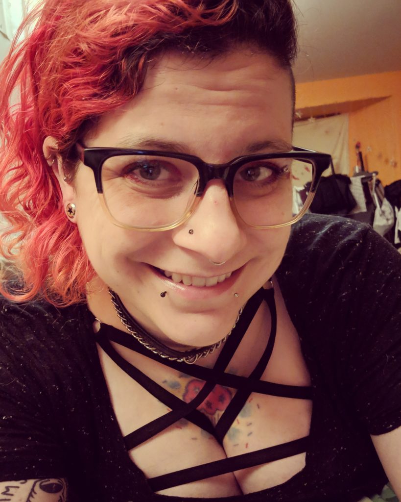 Image description: A queer white person is shown from the bust up, smiling into the camera. They have red hair and are wearing glasses, a strappy black top, and a chain maille necklace.