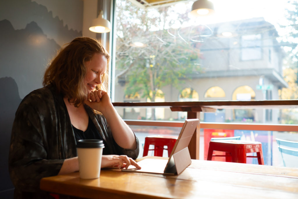 Body Positive and Fat Positive Business Branding Sessions for Podcasts, Authors, Writers, Photographers, and Dreamers
Author Thekla Richter is shown in a coffee shop with bright orange stools and a gray mural, working on a tablet or laptop with a cup of coffee in front of a large window showing a street scene. She is a plus-size white woman with red hair and is wearing a black top and gray cardigan.