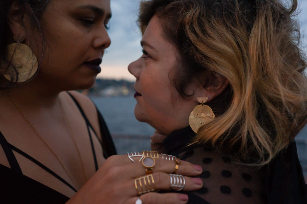 Body Positive and Fat Positive Product Photography Sessions for Products, Zines, Books, Art, Artwork, Crafts, Jewelry, Apparel, and Handmade Items
Plus-size models Croptopgoth and Adria Garcia stand close together in a sultry pose in front of Lake Union in Seattle, wearing rings and earrings made by Ohme Jewelry in gold and silver. One woman is of Native American descent and has long black hair. The other is a fat white woman with shoulder-length blonde hair.