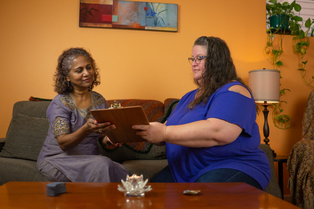 Body Positive and Fat Positive Business Branding Sessions for Massage Therapists, Energy Healers, Tarot Reading, Alternative Health, Meditation, Offices, Retail Locations, and Brick and Mortar
Two women sit on a gray couch in front of a brown coffee table and discuss an upcoming treatment in a massage therapy waiting room with orange walls. One woman is a fat white woman with long brown hair, wearing a blue top and jeans. The other is a woman of Indian heritage with shoulder-length brown and gray hair, wearing a lavender dress with gold detailing.