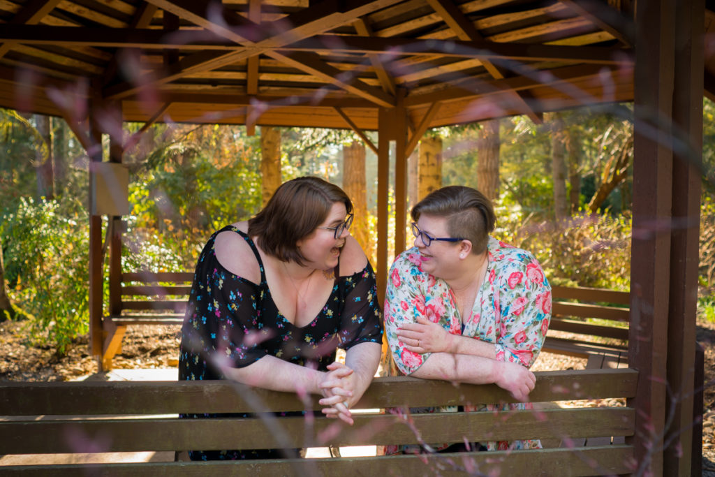 Body Positive and Fat Positive Business Branding Sessions for Podcasts, Authors, Writers, Photographers, and Dreamers
Eron Huenefeld and Amanda Underwood of the podcast Fat Face Feelings lean on a wooden railing and laugh under a large wooden gazebo on a bright day in a park with trees all around. Both are fat white women with glasses, short hair, and floral short-sleeve tops.