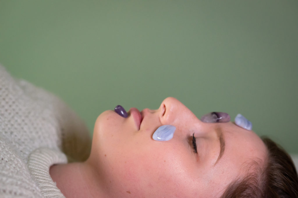 A young plus-size white woman lies on her back in front of a green wall, with her eyes closed. She has brown hair and is wearing a white sweater. Blue lace agate and amethyst tumbled gemstones have been placed on her forehead, chin and cheeks.
