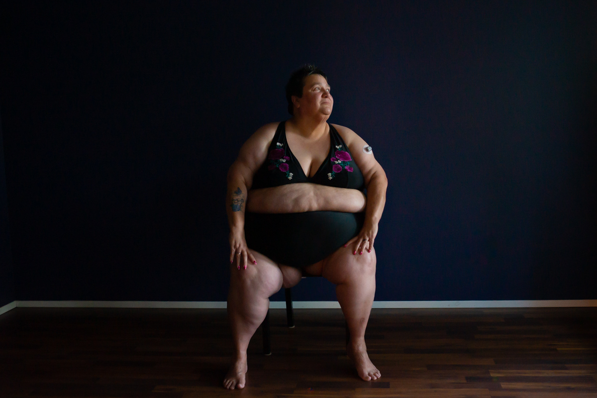 A fat woman with pale skin sits on a chair in a dark room, body facing forward and face turned to the right. She is wearing a black bikini with a floral design.